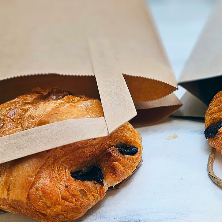 A close-up of a golden-brown flaky croissant partially tucked inside a plain brown paper bag on a light surface.