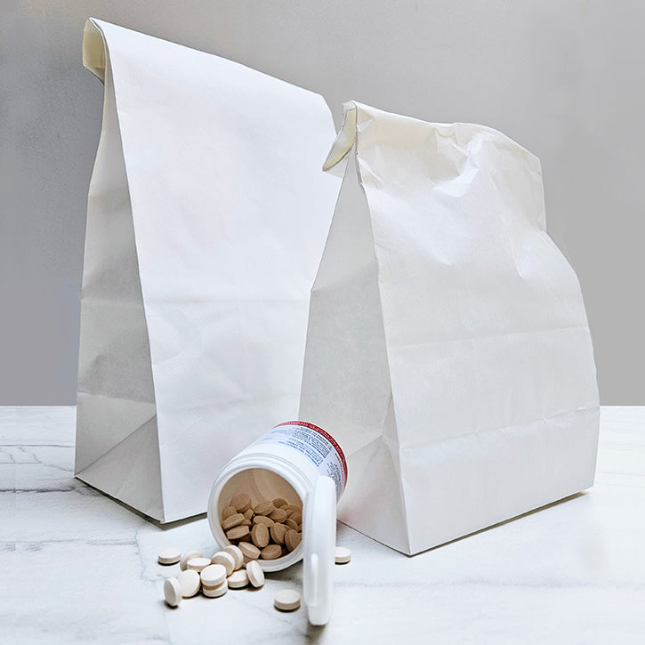 Two white paper pharmacy bags with a white pill bottle tipped over with beige tablets spilled onto a light marble surface against a neutral grey background.