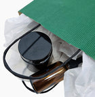 A close-up of a green paper gift bag with white tissue paper, revealing a black jar and an amber glass cosmetics bottle.
