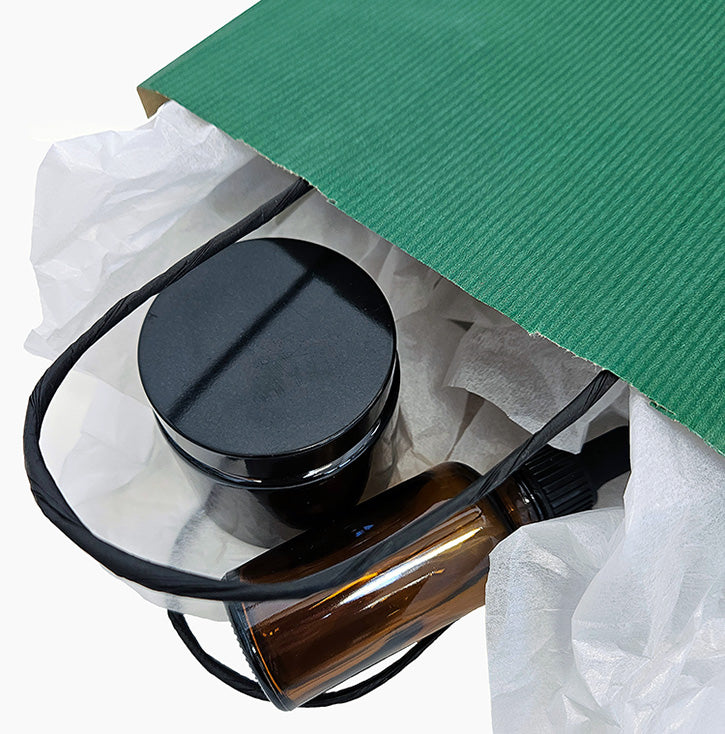 A close-up of a green paper gift bag with white tissue paper, revealing a black jar and an amber glass cosmetics bottle.