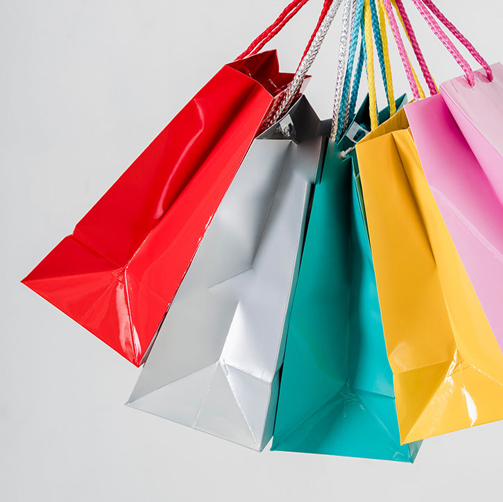 A cluster of glossy paper shopping bags in red, white, teal, yellow, and pink, hanging against a light grey background.